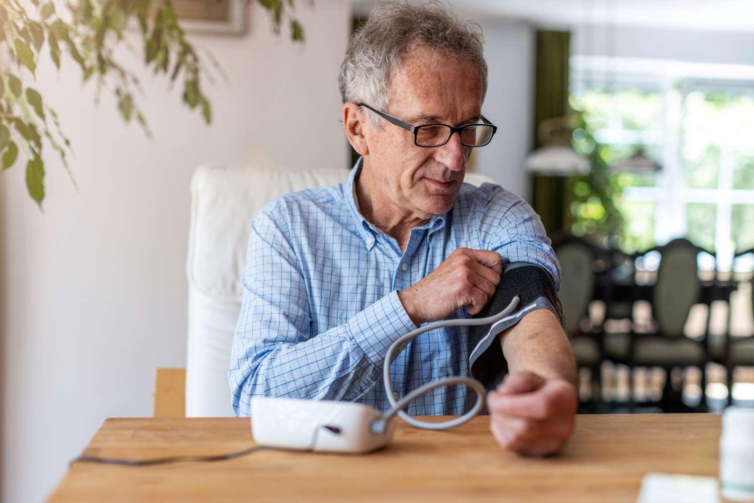 Blutdruckmessgeräte: Älterer Mann sitzt an einem Tisch und befestigt ein Blutdruckmessgerät an seinem Arm.