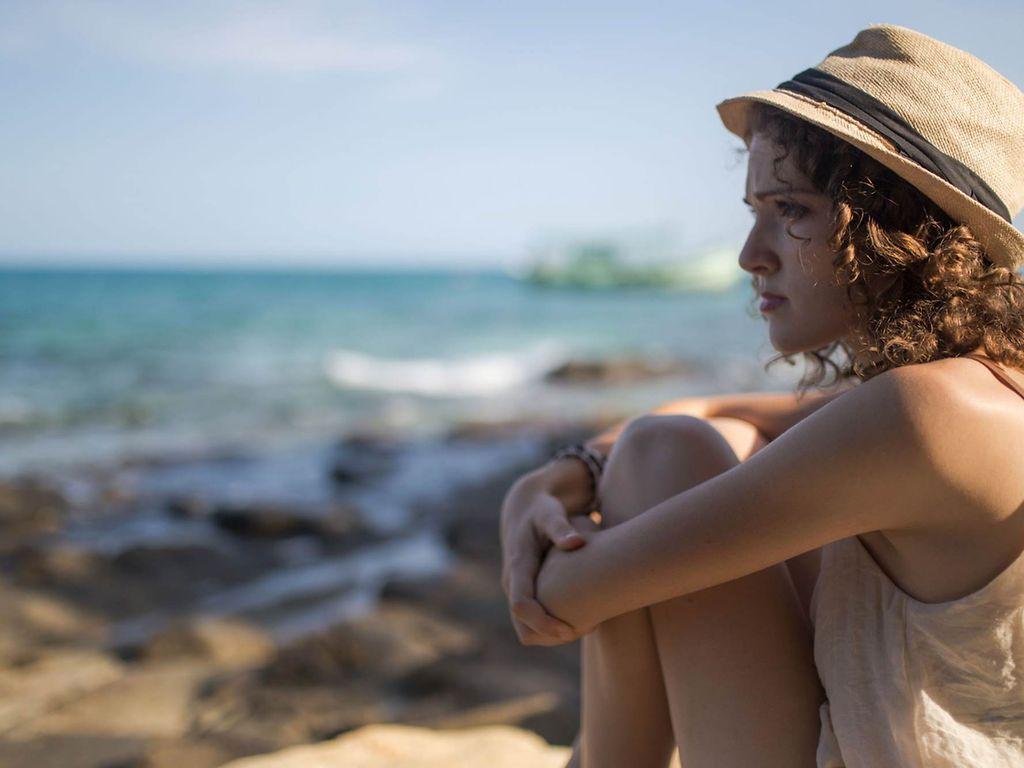 Sonnenallergie: Frau mit Hut und ernstem Gesichtsausdruck sitzt am Wasser.