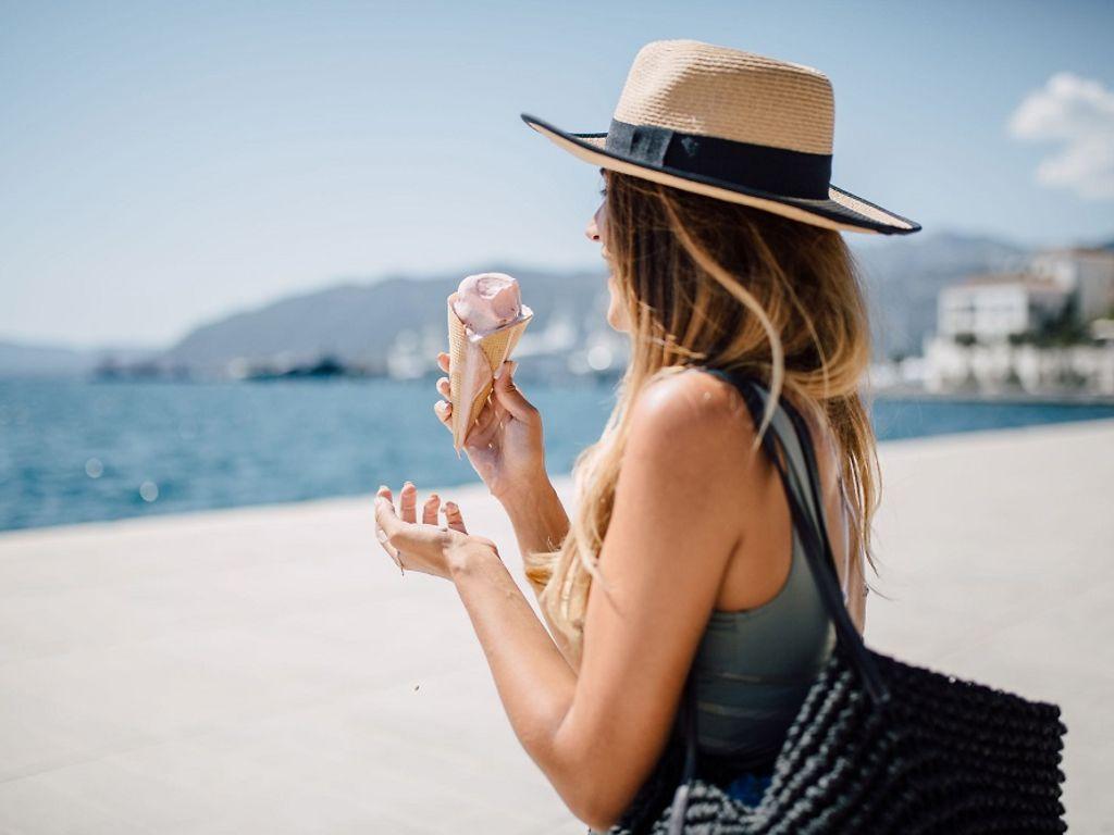 Sonnenstich: Frau mit Sonnenhut isst ein Eis am Strand.