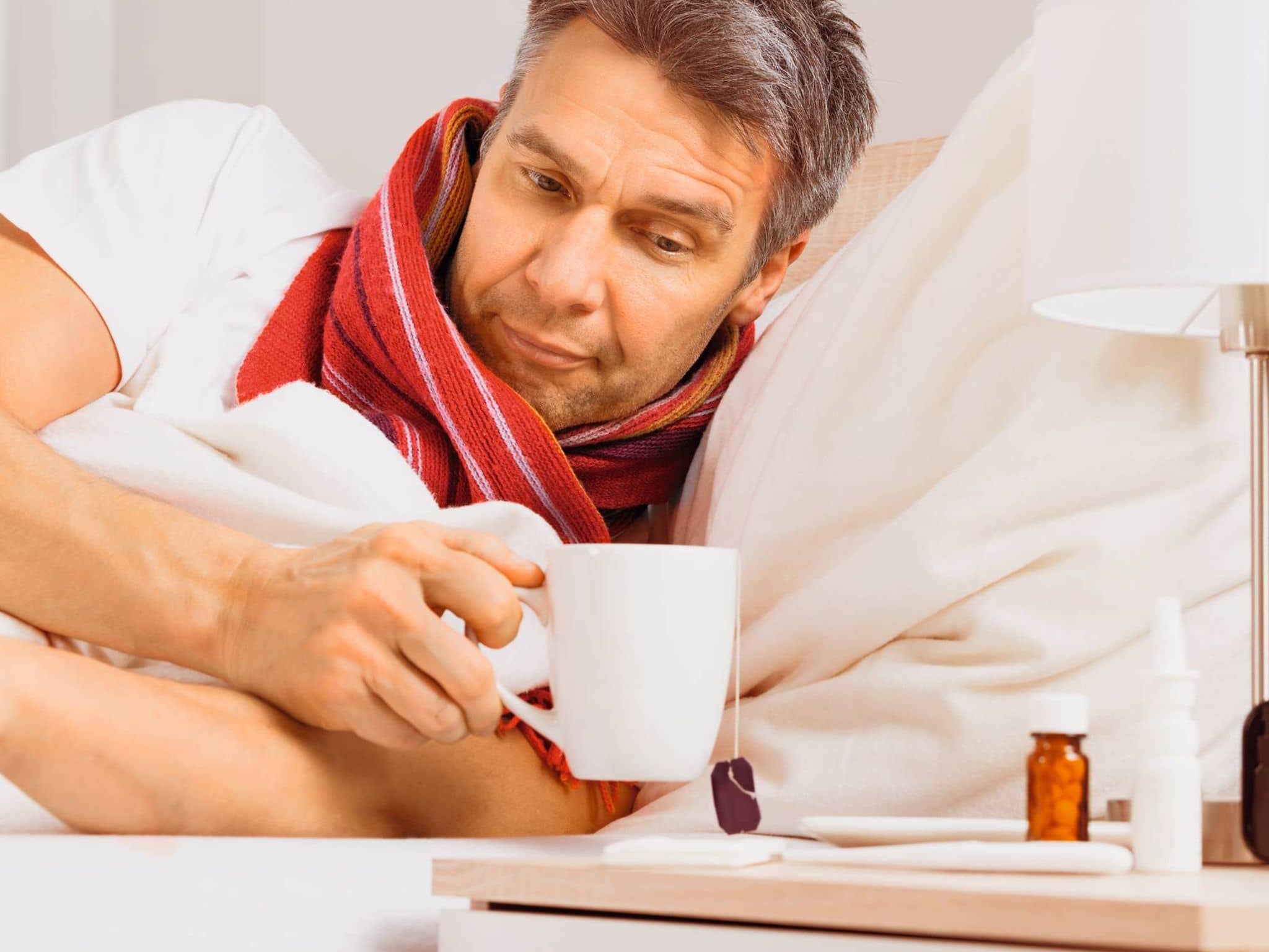 Ein Mann liegt mit Schal im Bett und trinkt Tee - ein bewährtes Hausmittel gegen Halsschmerzen