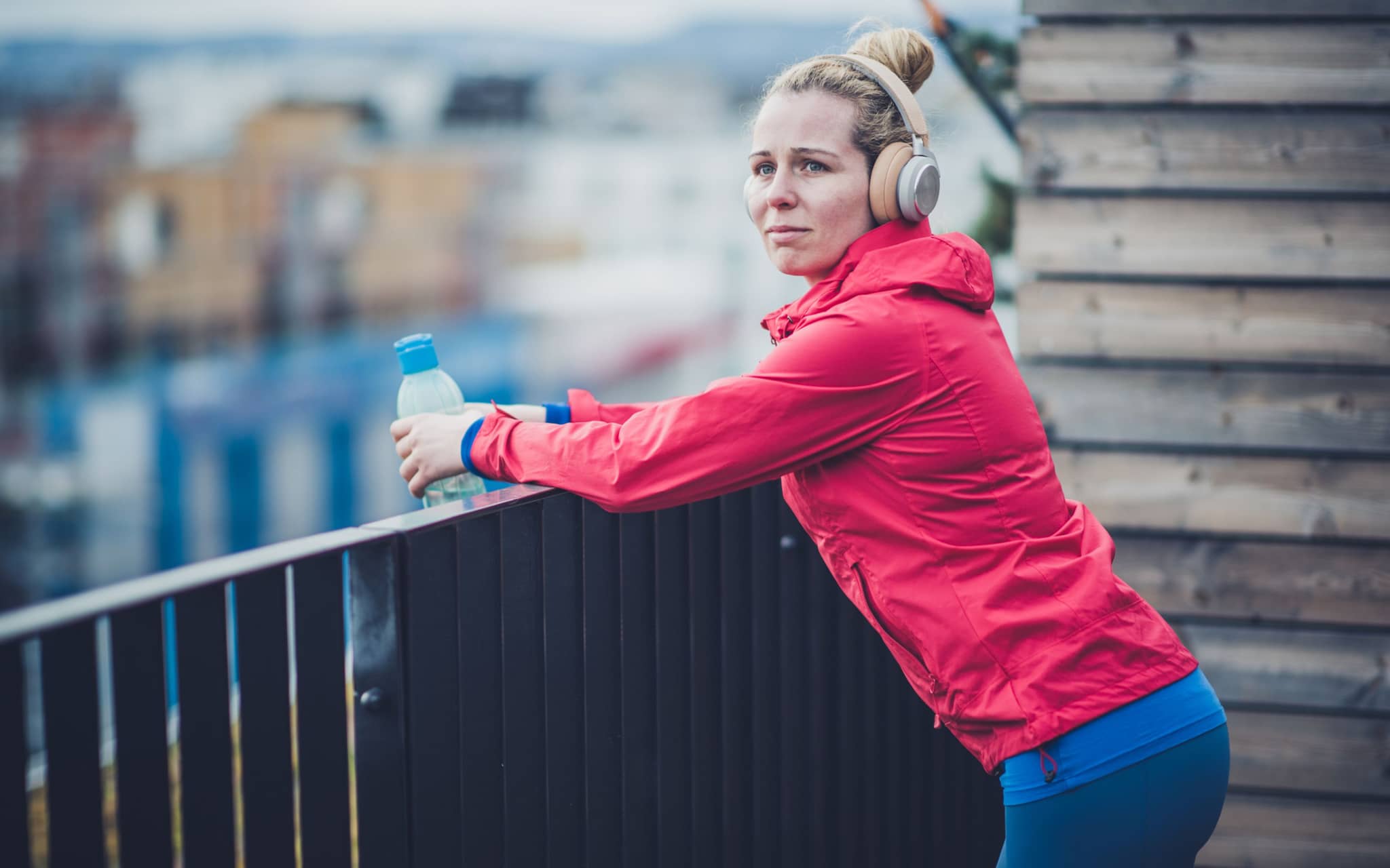 Eine junge Frau in Sportkleidung hört Musik und lehnt an einer Balkonbrüstung.