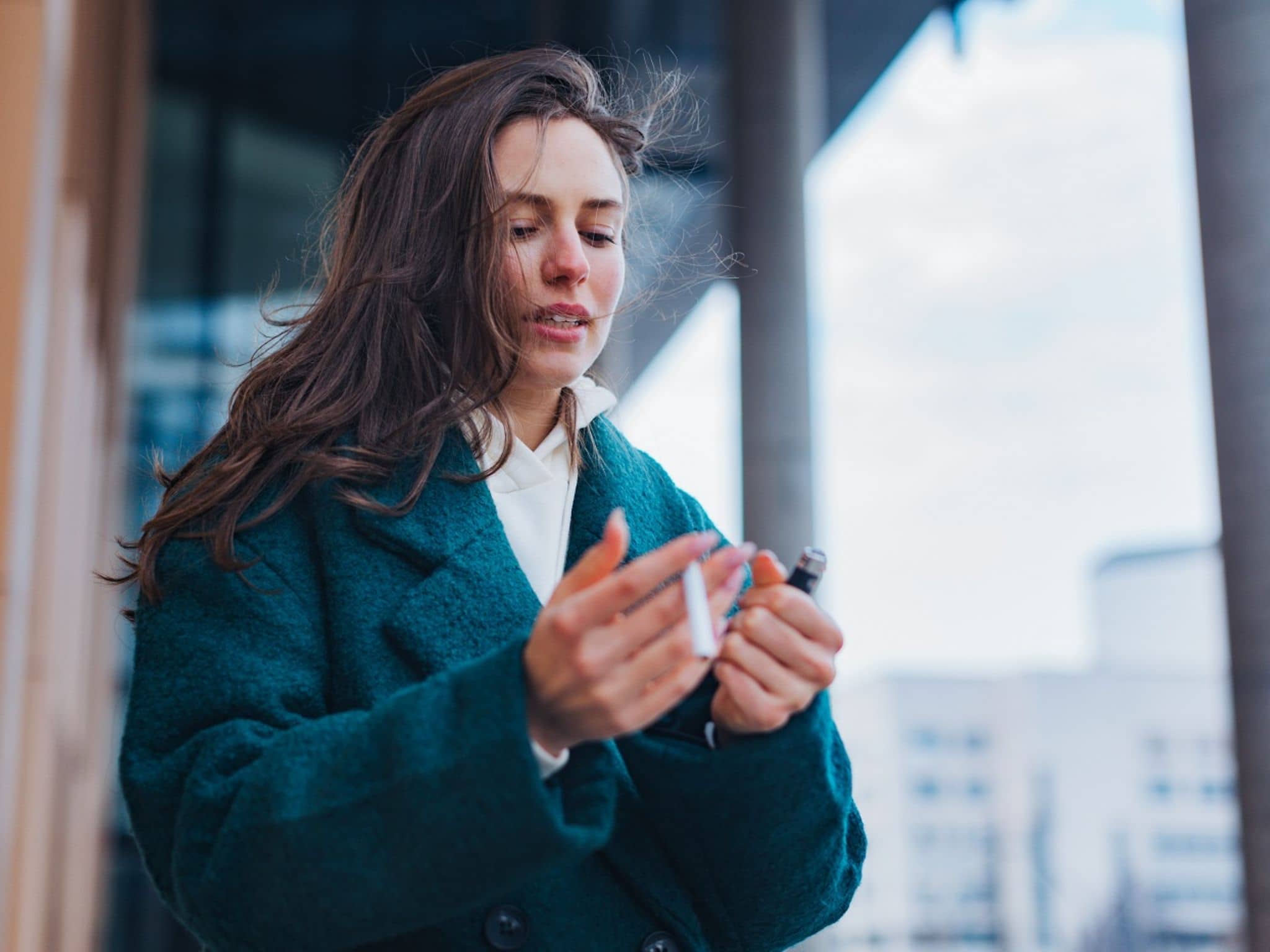 Eine junge Frau steht draußen mit einer Zigarette und einem Feuerzeug in der Hand und guckt auf ihre Hände.