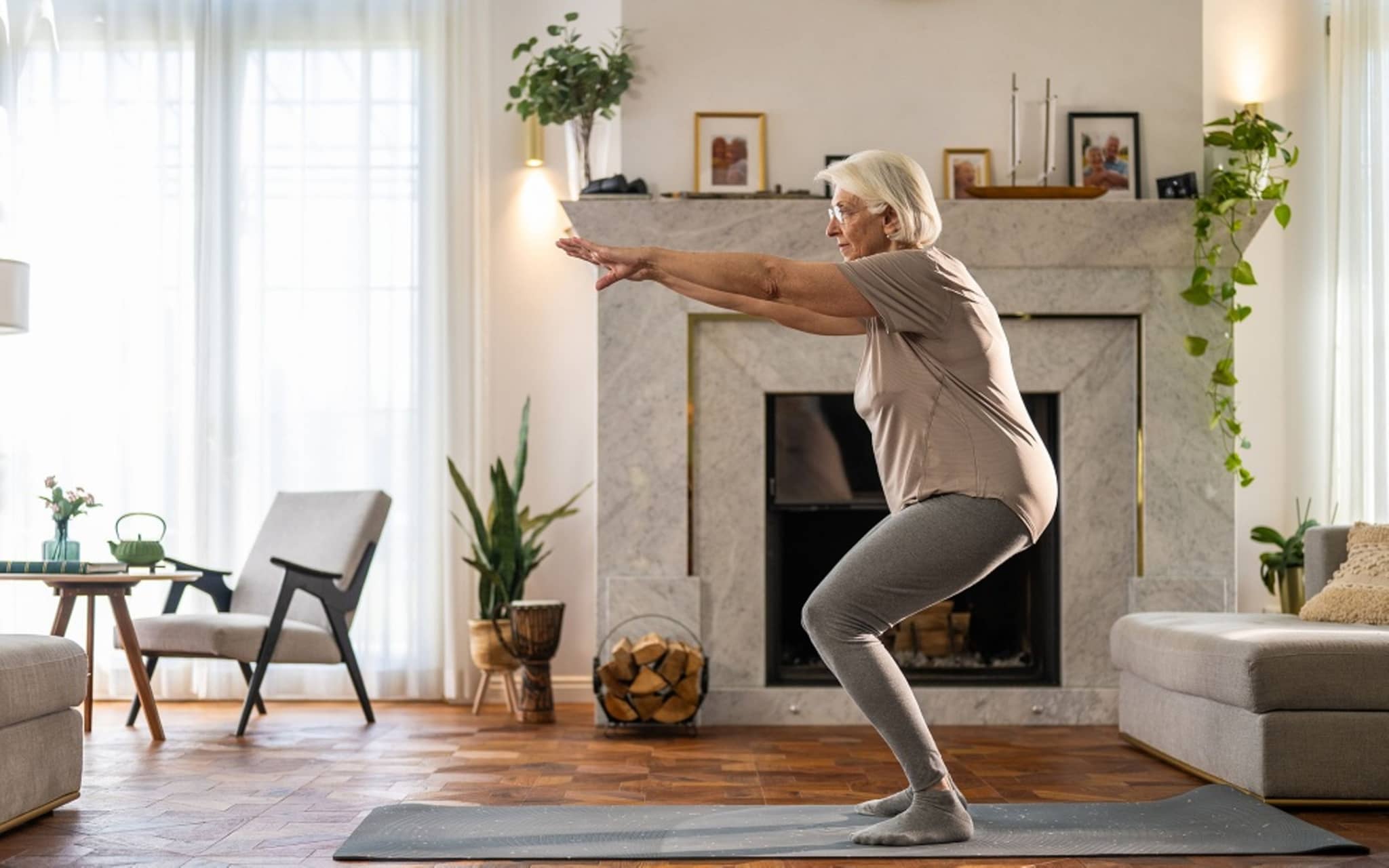 Ältere Frau macht eine Kniebeuge auf einer Yogamatte im Wohnzimmer.