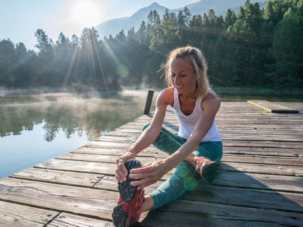 Laufen macht glücklich: Eine Frau sitzt auf einem Steg am See und stretcht sich.