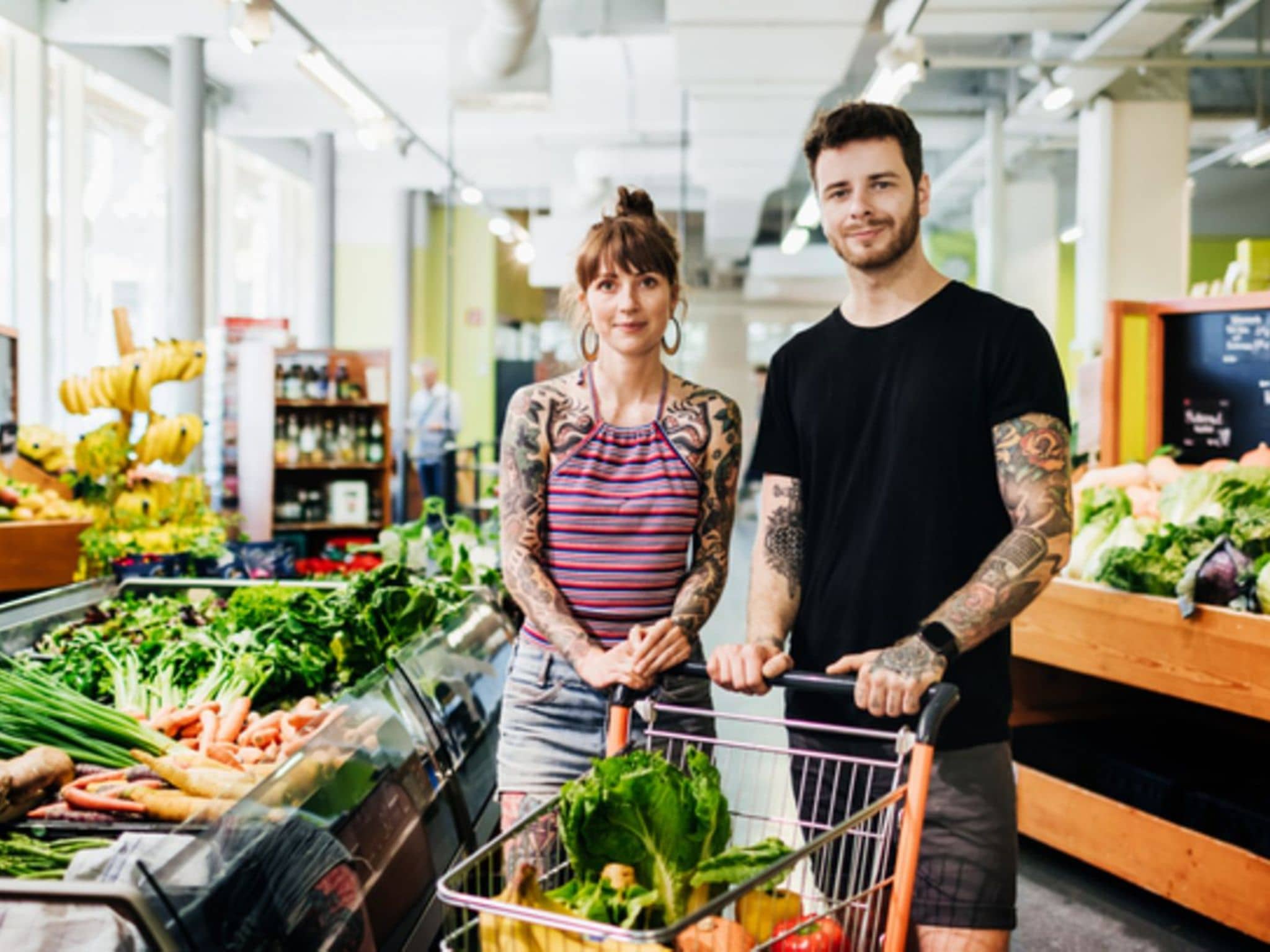 Vegane Ernährung und Nährstoffe: Junges Paar steht mit einem gefüllten Einkaufswagen in der Obst- und Gemüseabteilung.