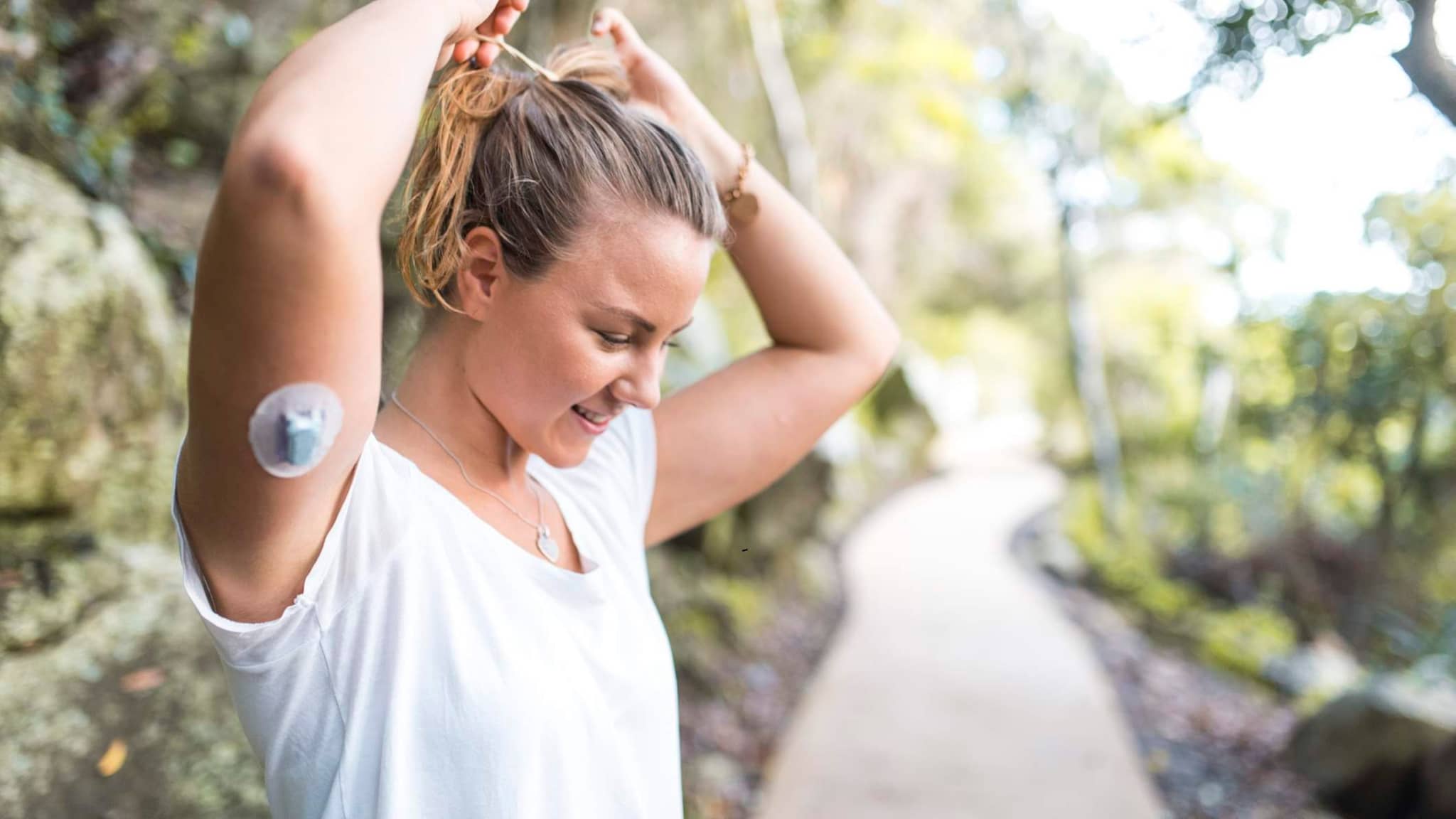 Diabetes: Frau mit Diabetessensor am Arm macht sich einen Zopf in der Natur.