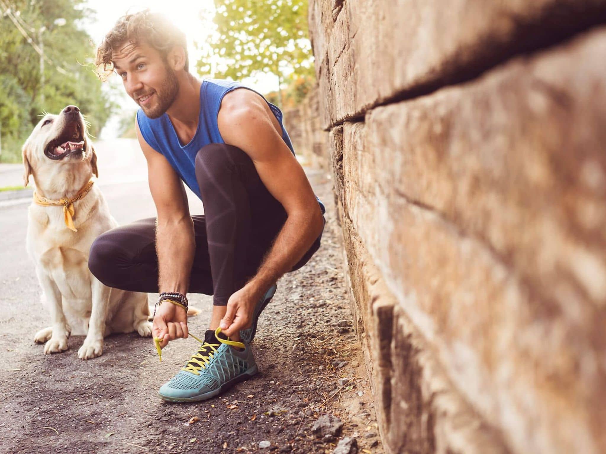 Schwitzender Mann macht sich kniend einen Laufschuh zu, daneben sitzt sein Hund.