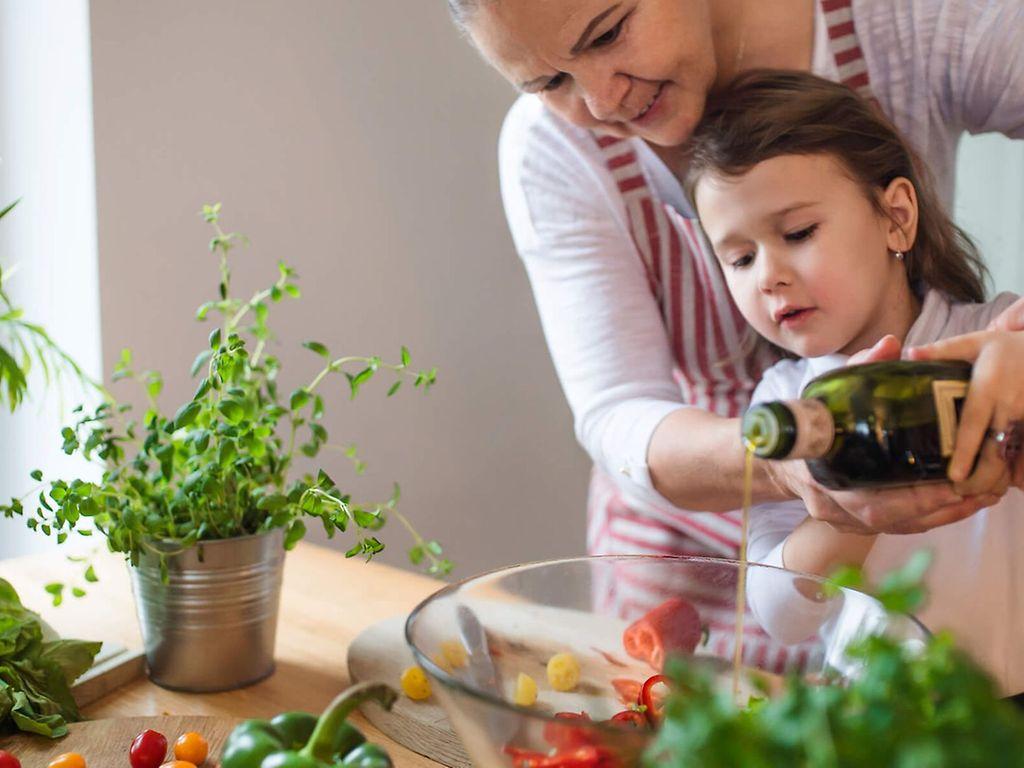 Makronährstoff Fett: Kleines Mädchen gießt gemeinsam mit seiner Mutter Öl über einen Salat.