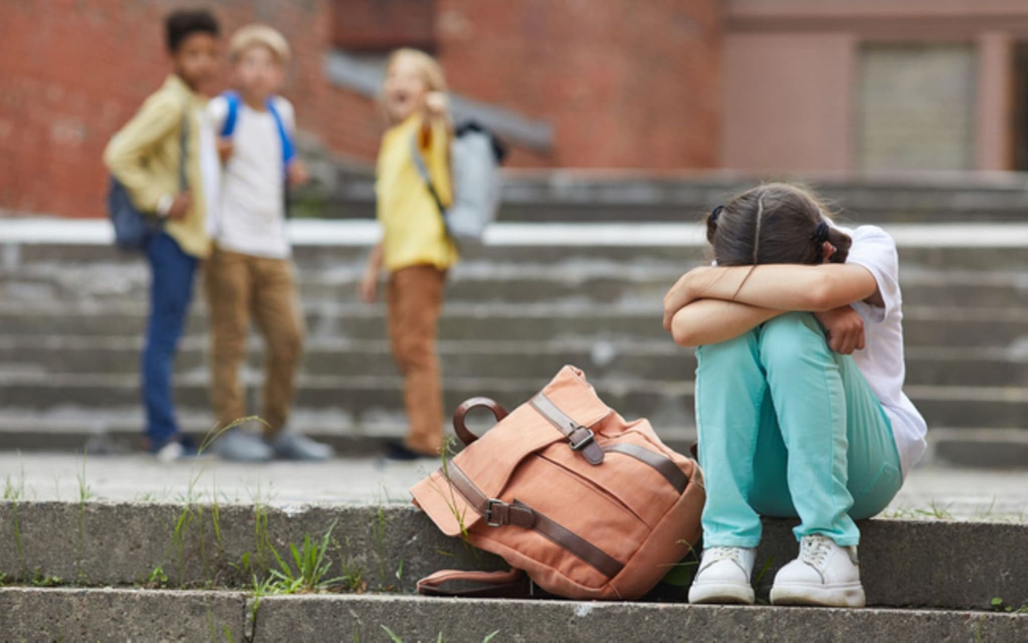 Mobbing: Ein Mädchen sitzt traurig auf dem Schulhof. Im Hintergrund sind mehrere Kinder, die auf sie zeigen.