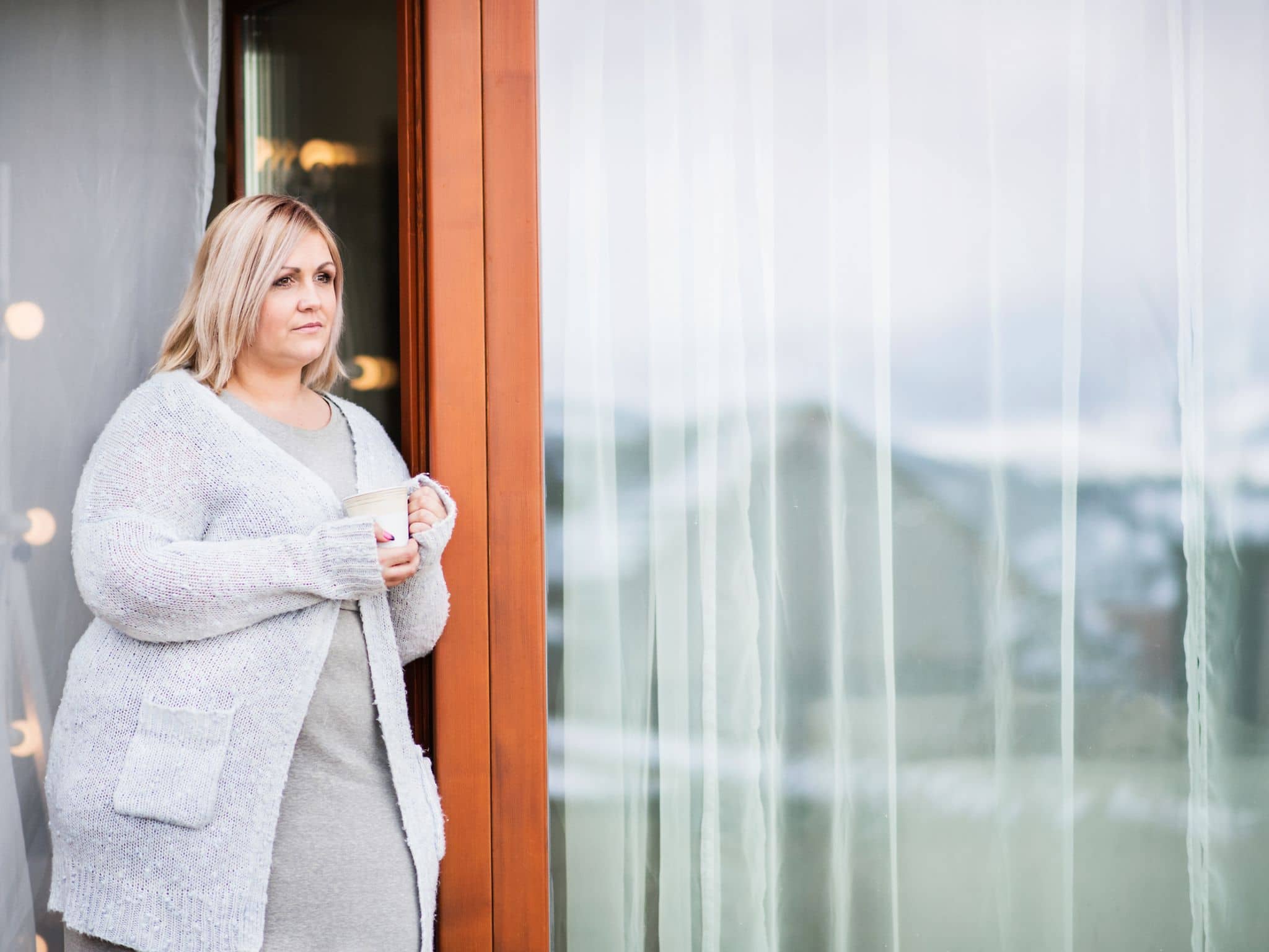 Adipositas: Übergewichtige Frau lehnt an einer Terrassentür und schaut traurig zur Seite.