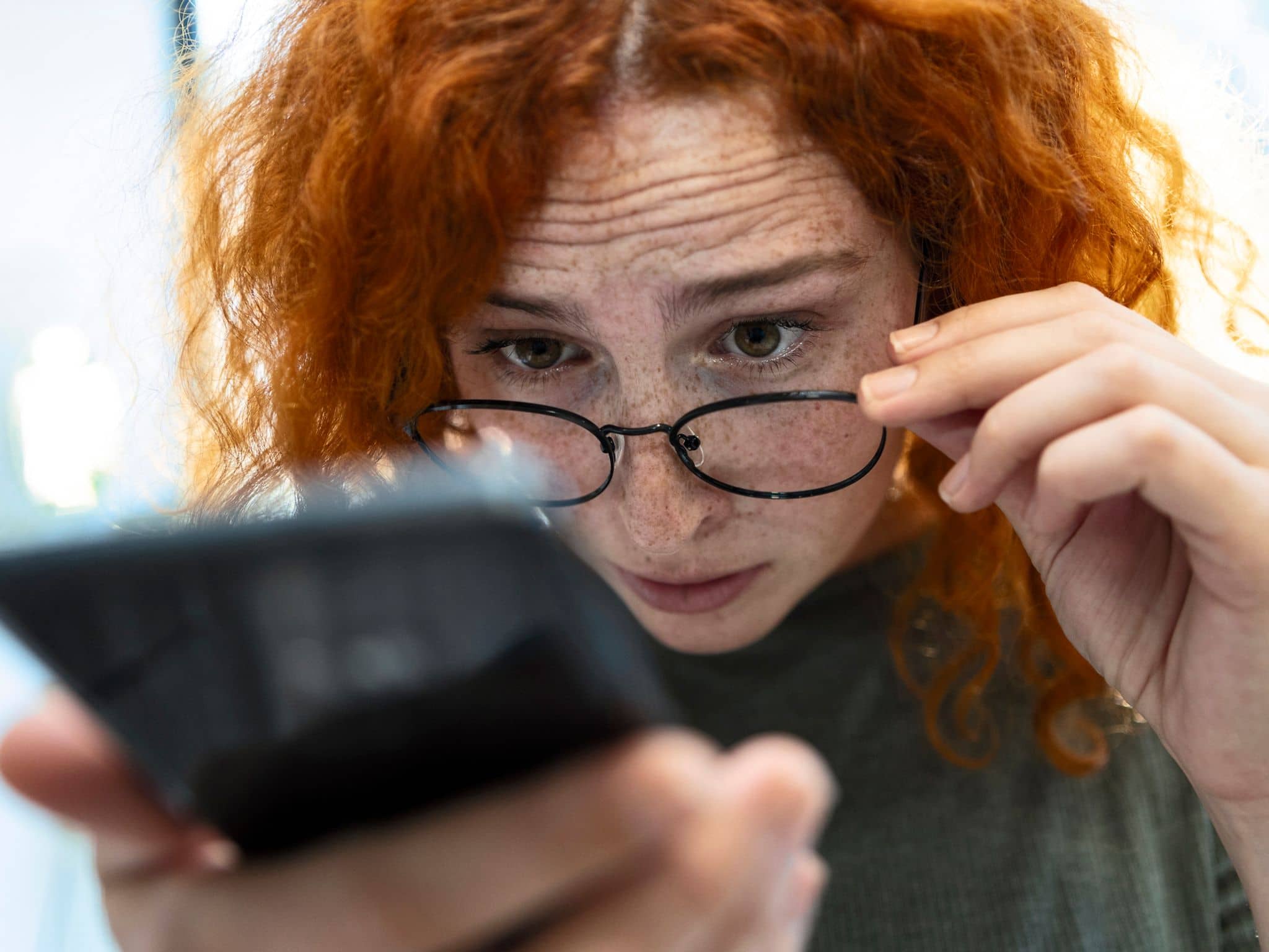 Frau schaut über ihre Brille auf ein Smartphone in ihrer Hand.