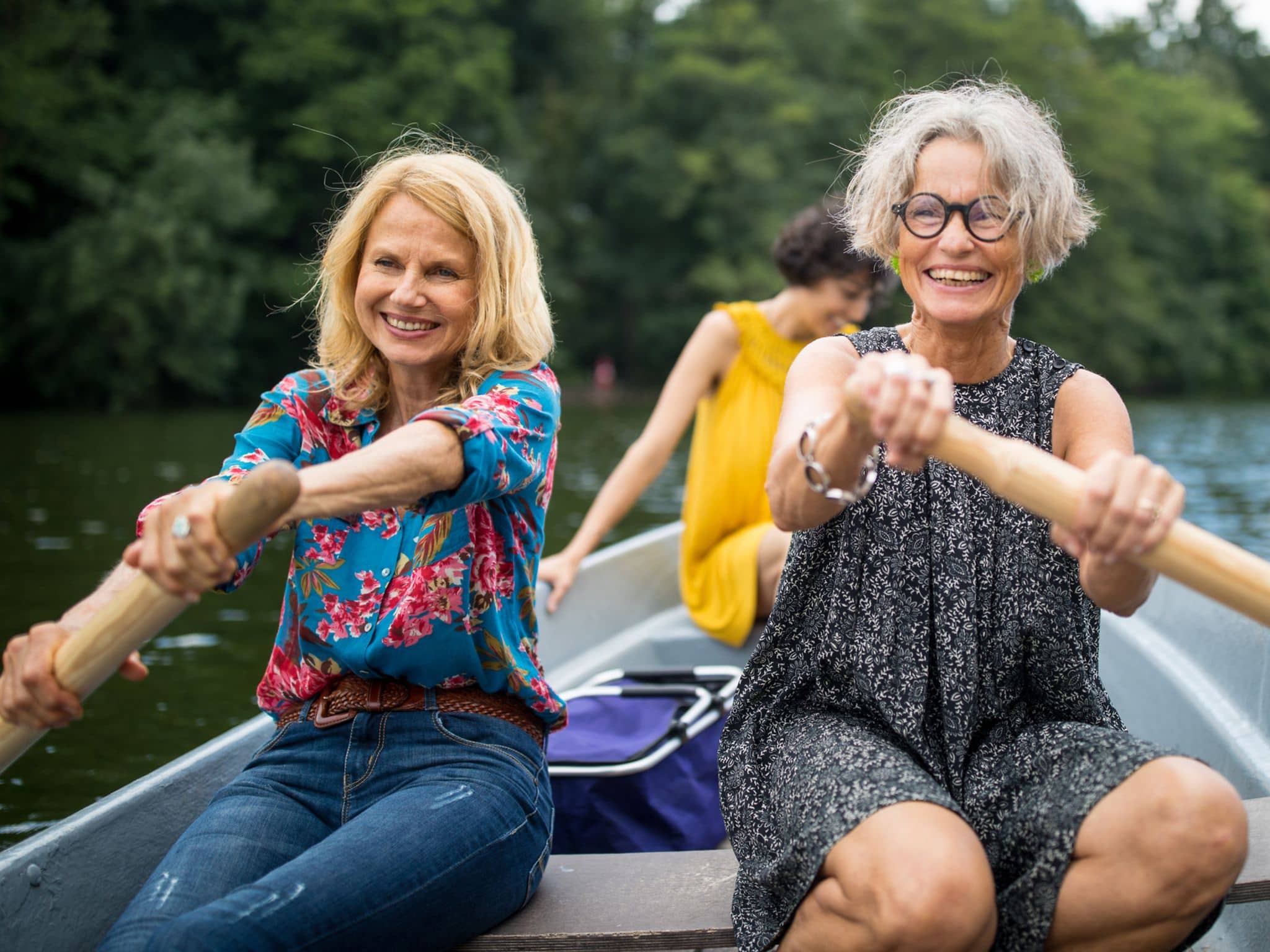 Zwei ältere Frauen rudern in einem Boot, mit einer dritten Frau im Hintergrund.