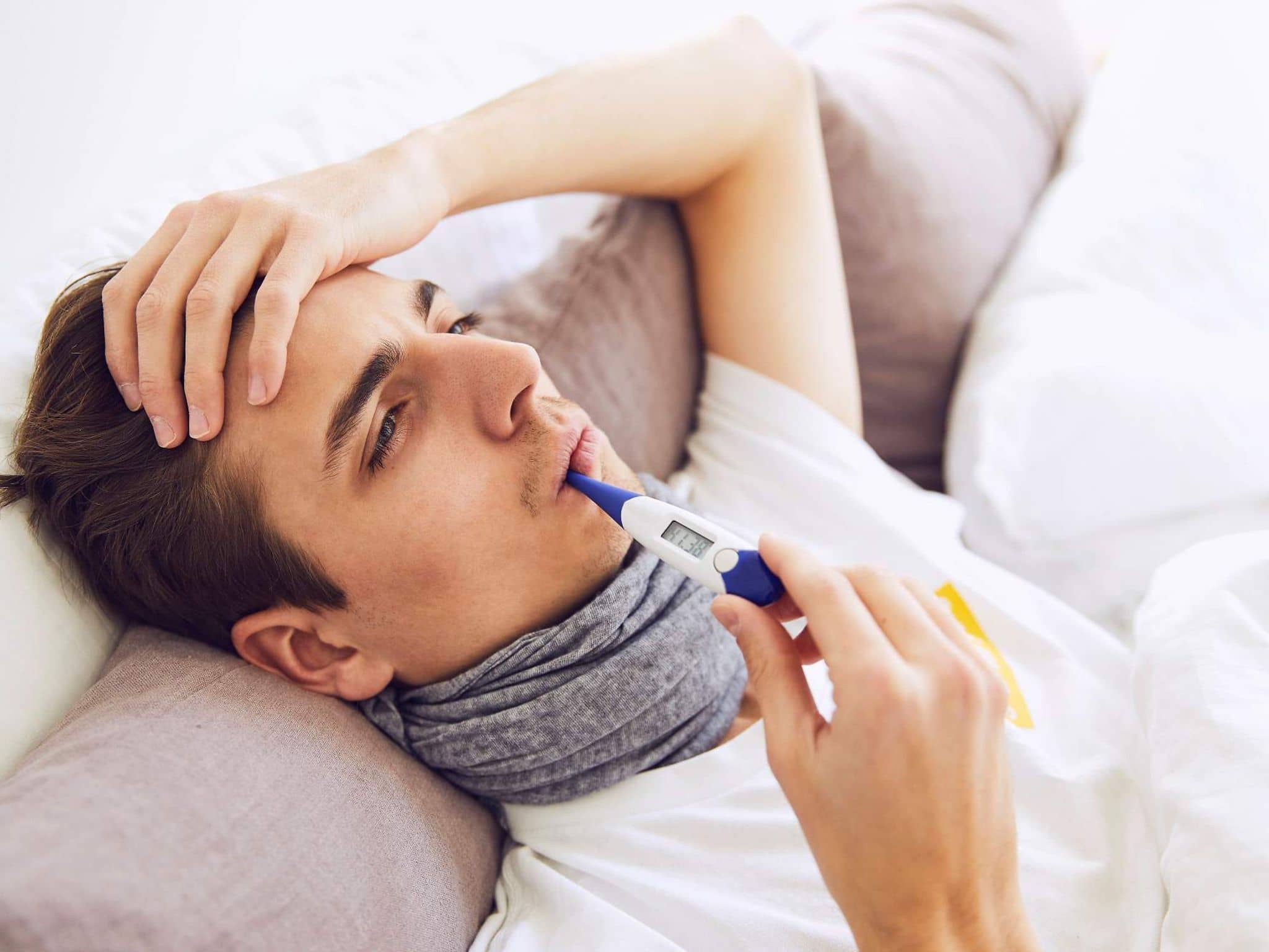 Lungenentzündung: Junger kranker Mann auf dem Sofa mit Halstuch hat Fieberthermometer im Mund und hält seine Hand an die Stirn.