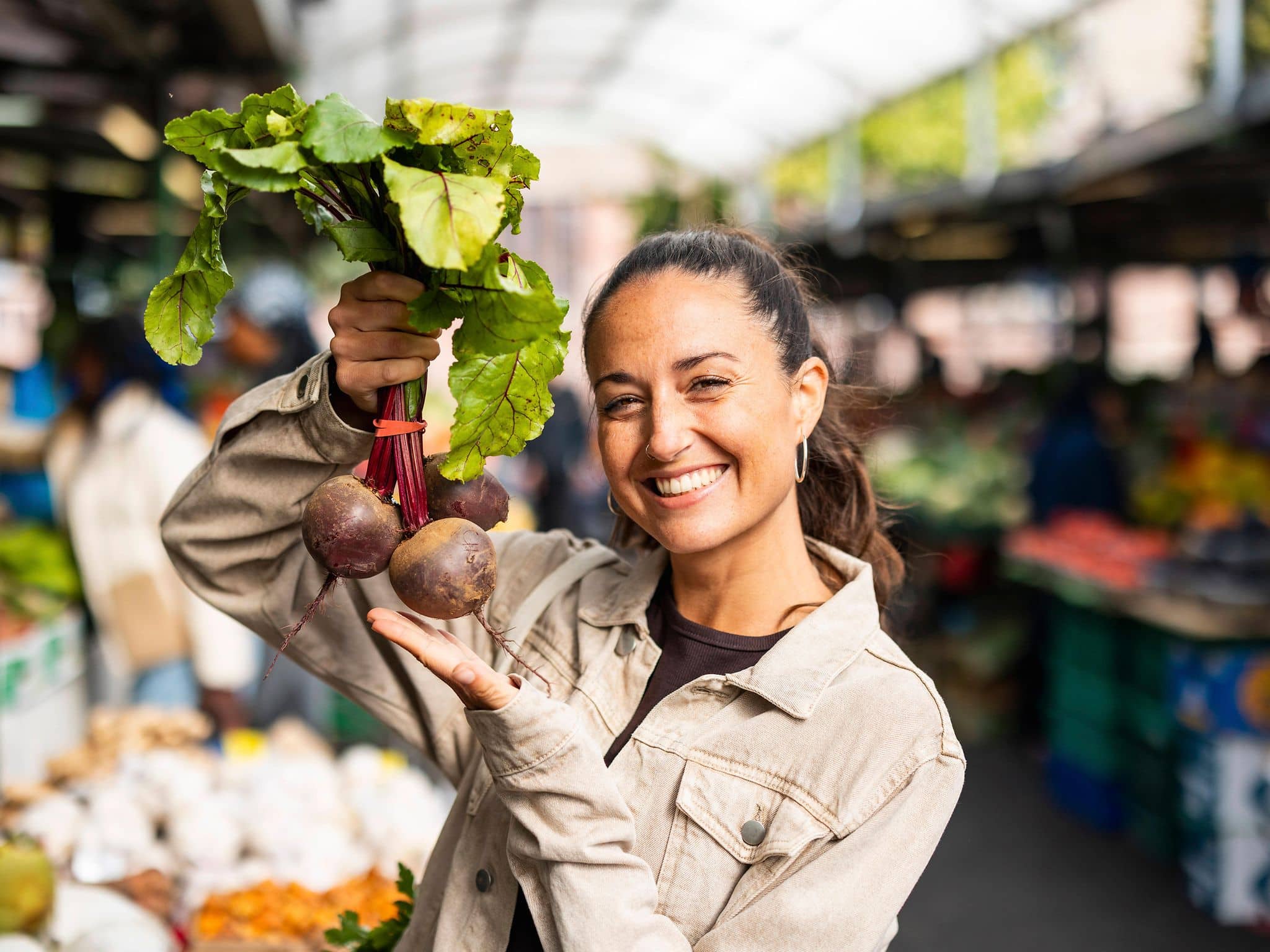 Frau hält lächelnd Rote Bete vor einem Marktstand in die Höhe.