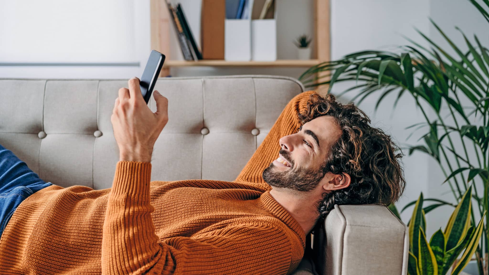 Newsletter bestellen: Lächelnder Mann liegt auf Couch und hält Smartphone in der Hand