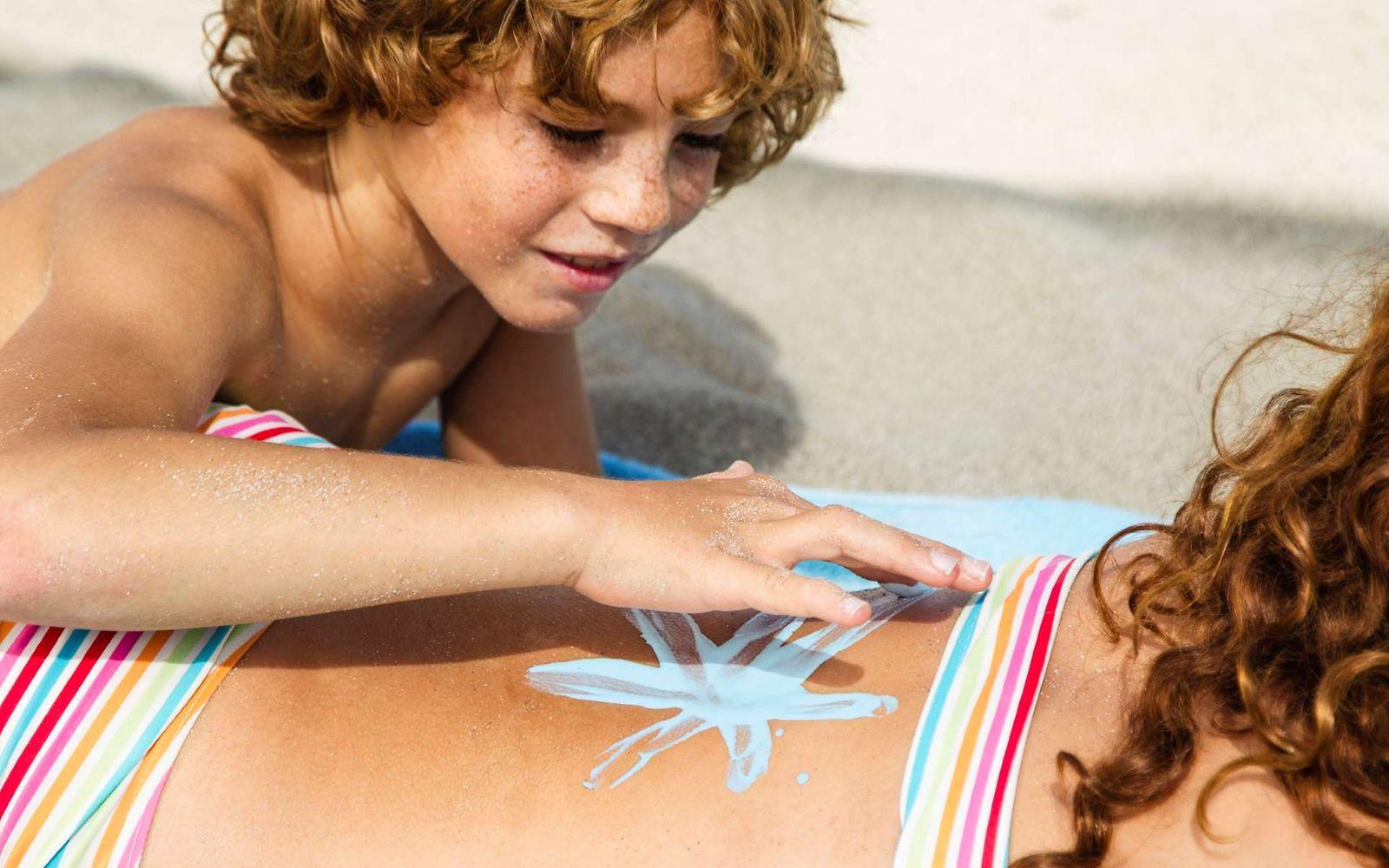 Ein Kind liegt am Strand und malt einer Frau mit Sonnencreme einen Stern auf den Rücken.