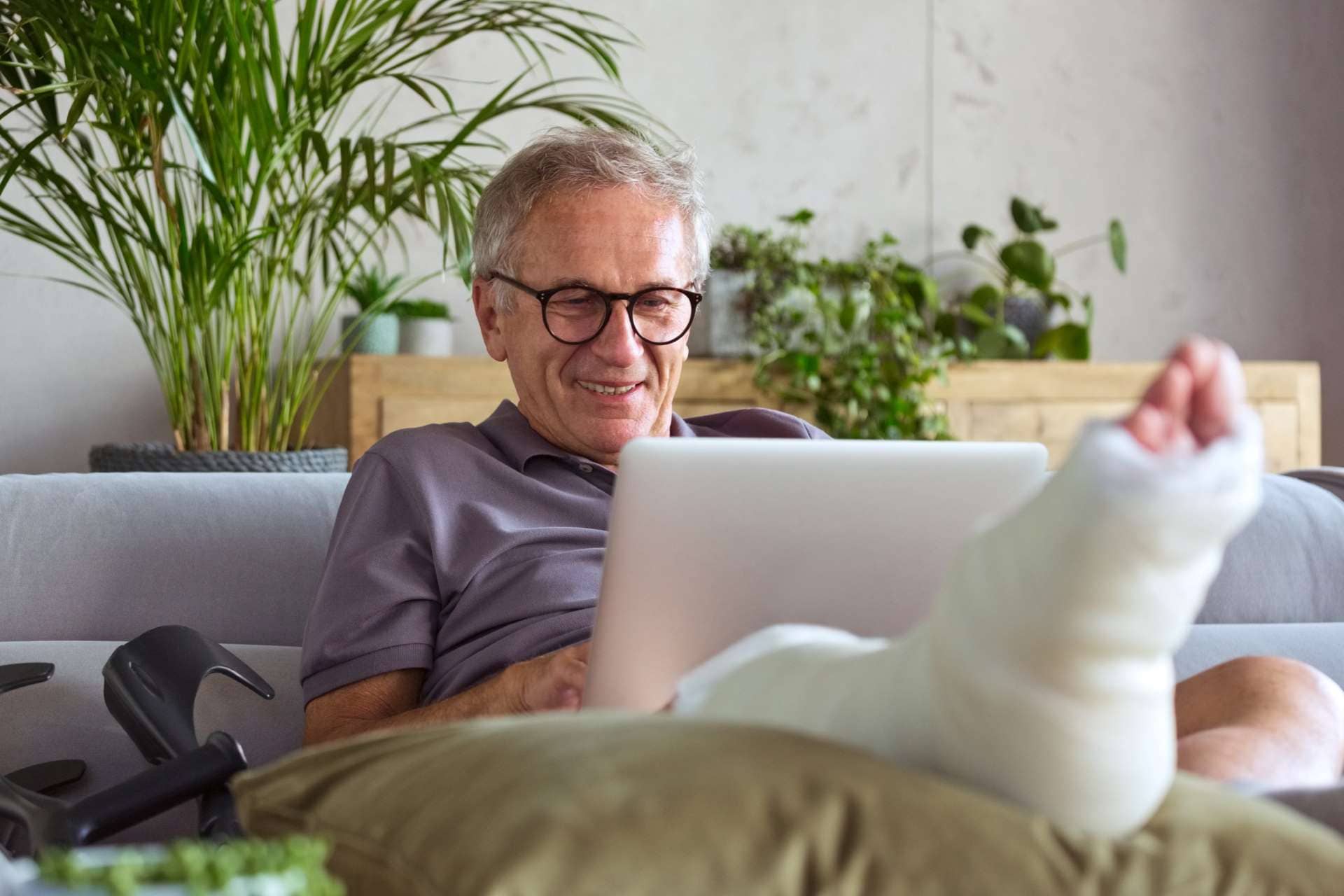 Älterer Mann mit Gips am Fuß sitzt auf dem Sofa und nutzt ein Tablet.