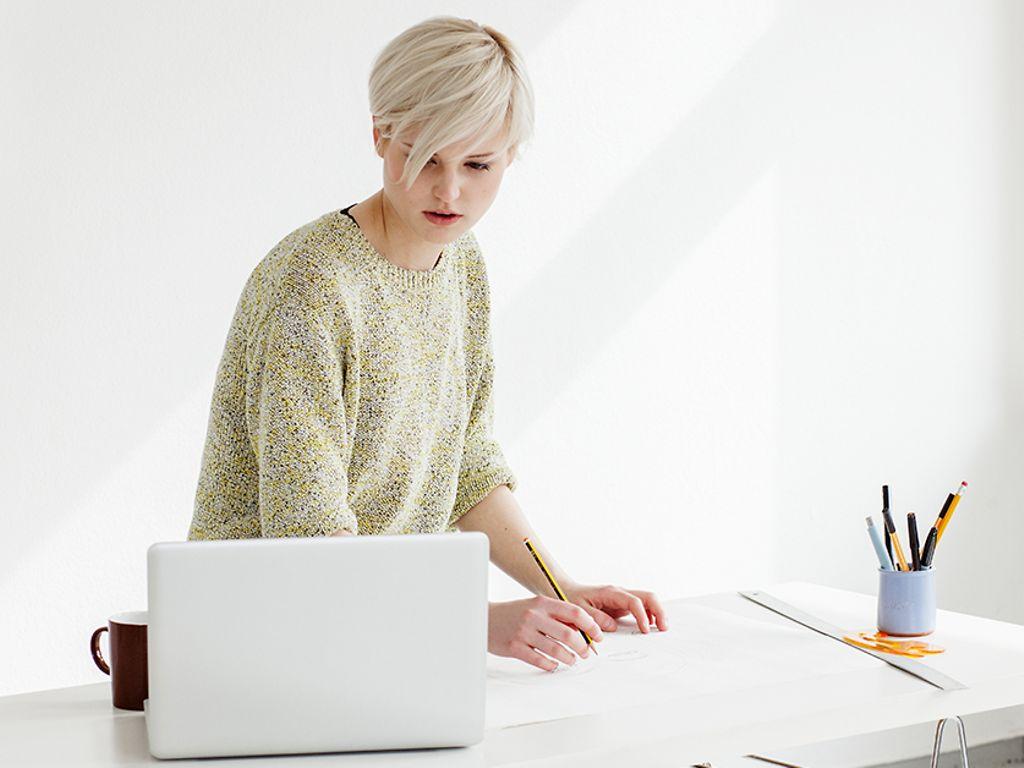 Junge Frau zeichnet konzentriert mit einem Bleistift und schaut auf ihre Vorlage auf dem Laptop