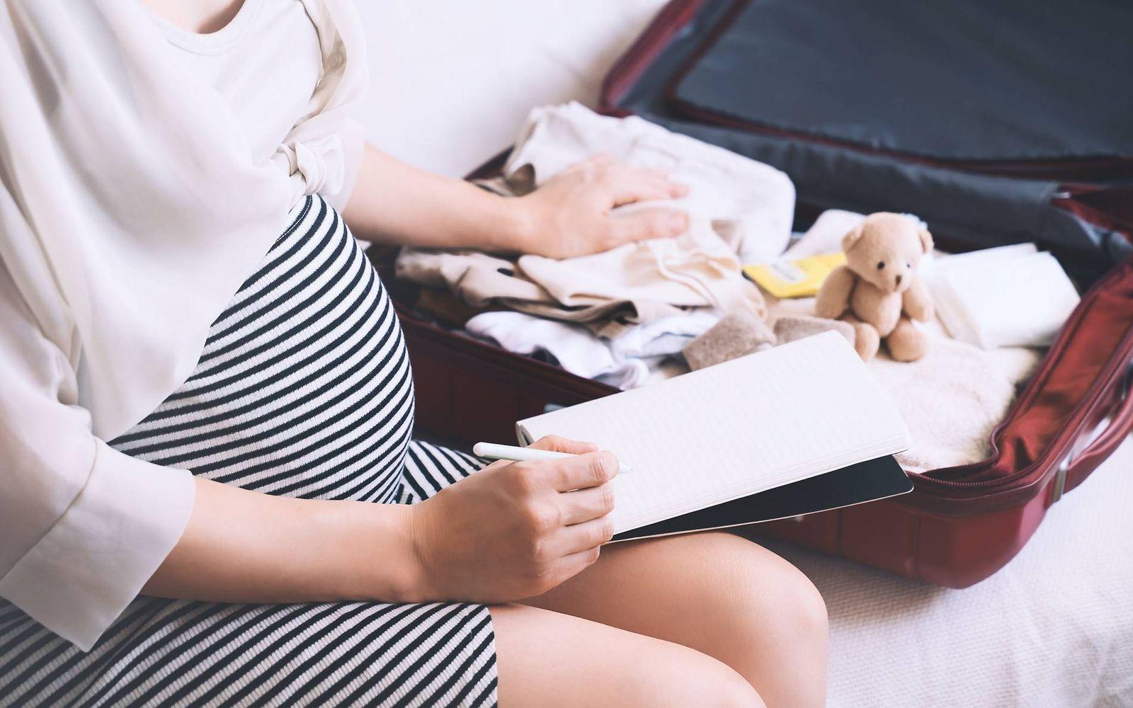 Kliniktasche packen: Schwangere hakt Liste ab, im Hintergrund ist ein gepackter Koffer zu sehen