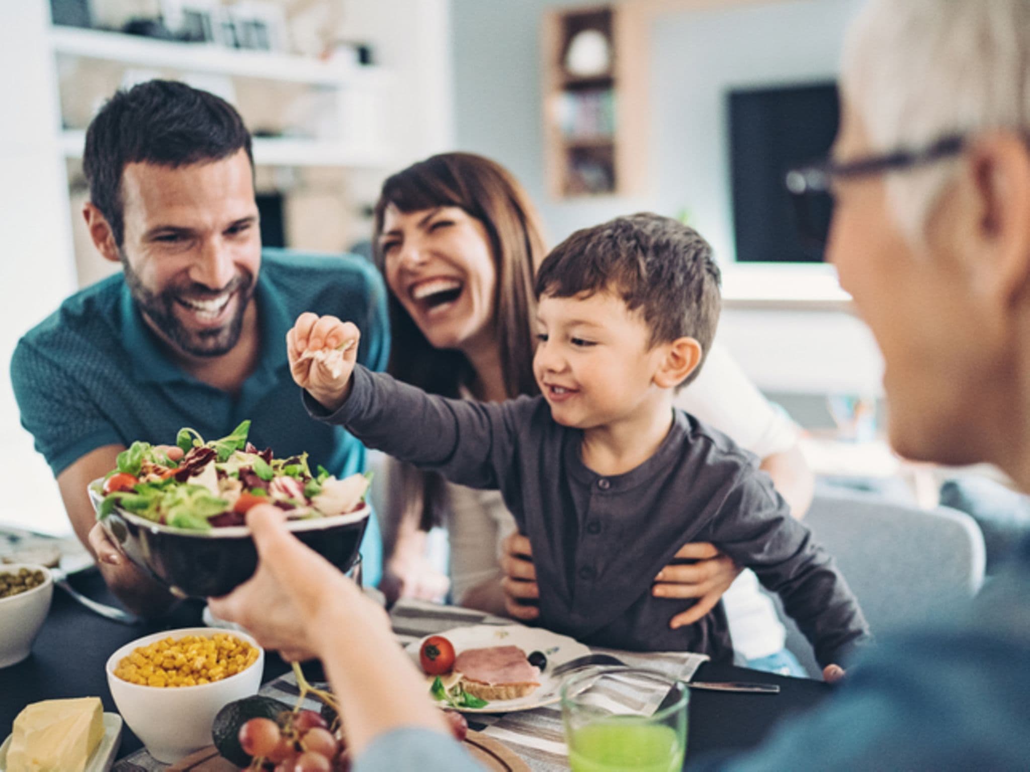 Ernährungstipps: Fröhliche Familie mit Kind sitzt am Tisch und isst Salat.