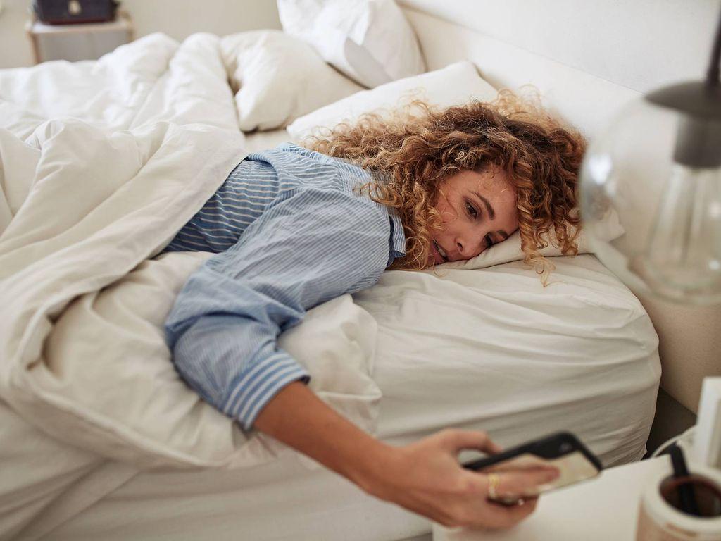 Morgenmuffel: Frau liegt im Bett und schaut müde auf ihr Handy