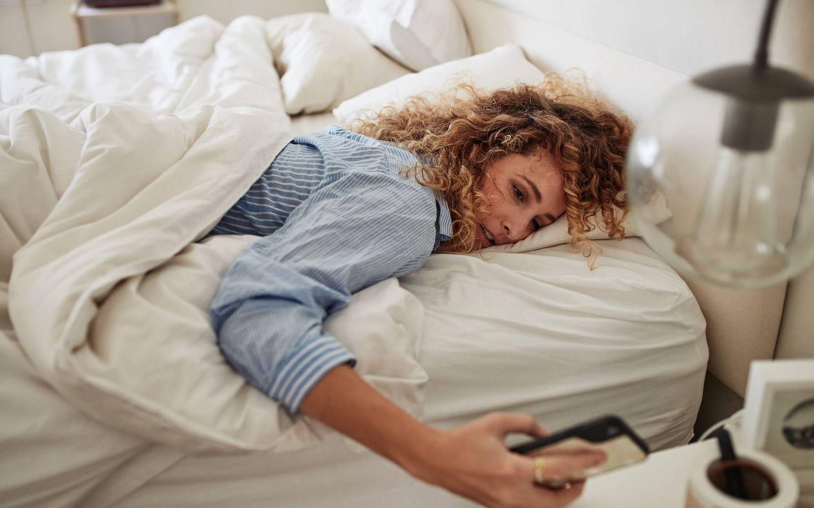 Morgenmuffel: Frau liegt im Bett und schaut müde auf ihr Handy