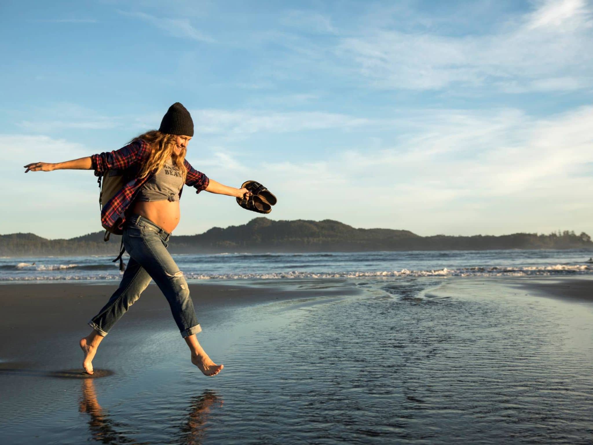Eine junge Schwangere spaziert fröhlich barfuß am Strand.