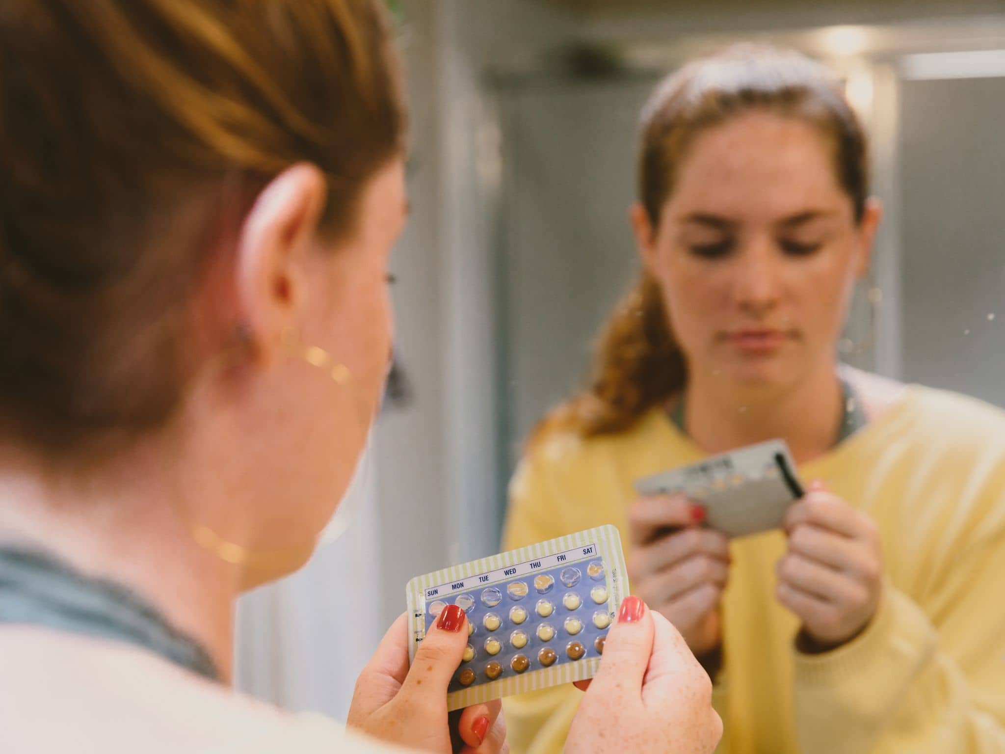 Symbolbild Pillenarten: Eine junge Frau steht vor einem Spiegel im Badezimmer und betrachtet eine Pillenpackung