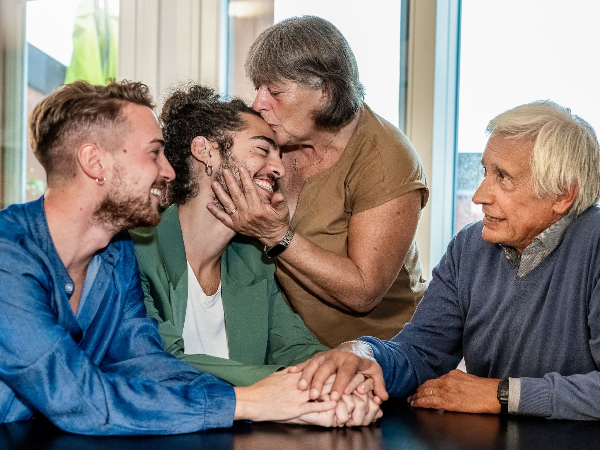 Symbolbild für Outing: Zwei Männer sitzen am Esstisch bei den Eltern, die Mutter küsst einen der Männer liebevoll auf die Stirn