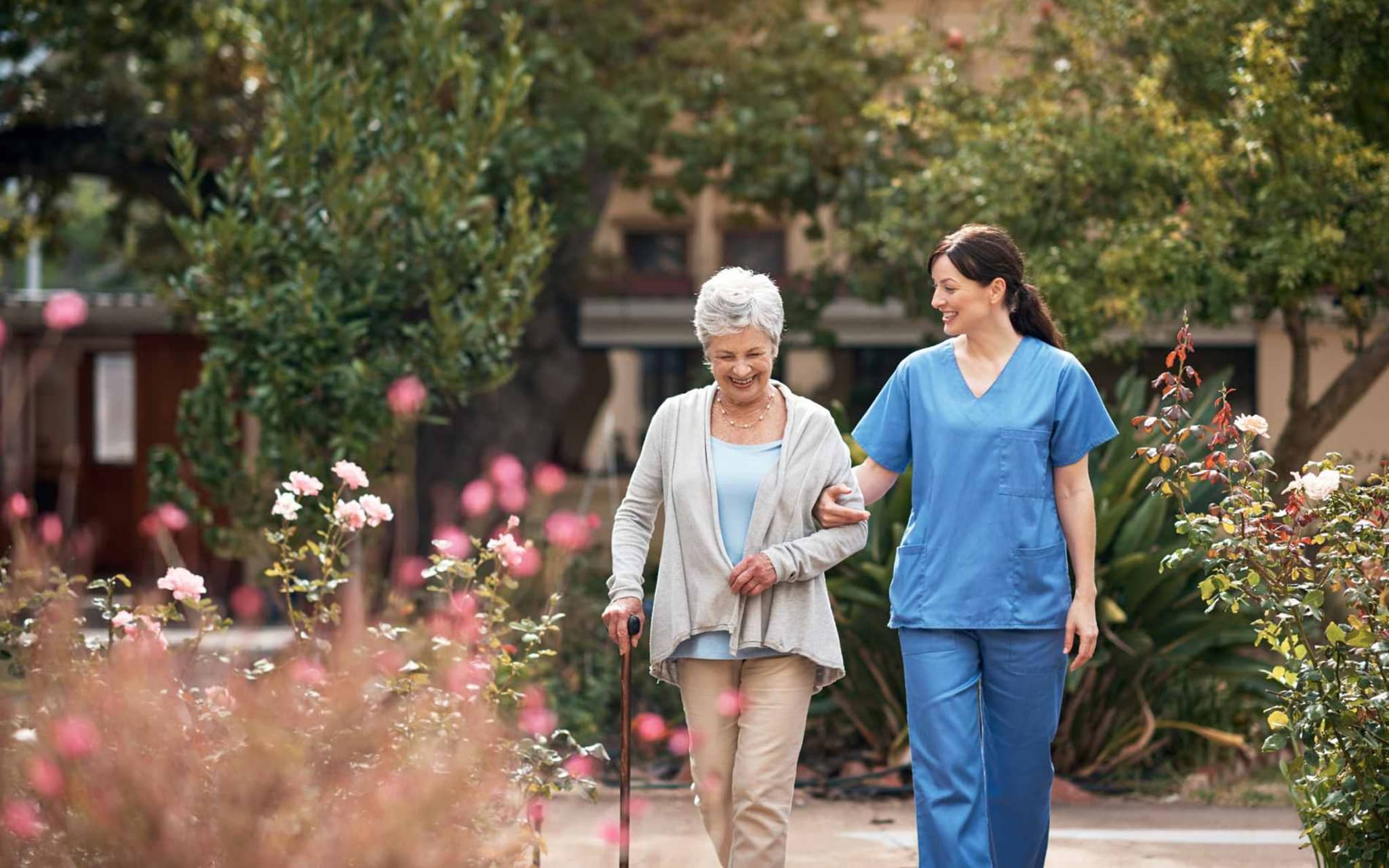 Bild: Pflegerin und Patientin gehen im Rosengarten spazieren.