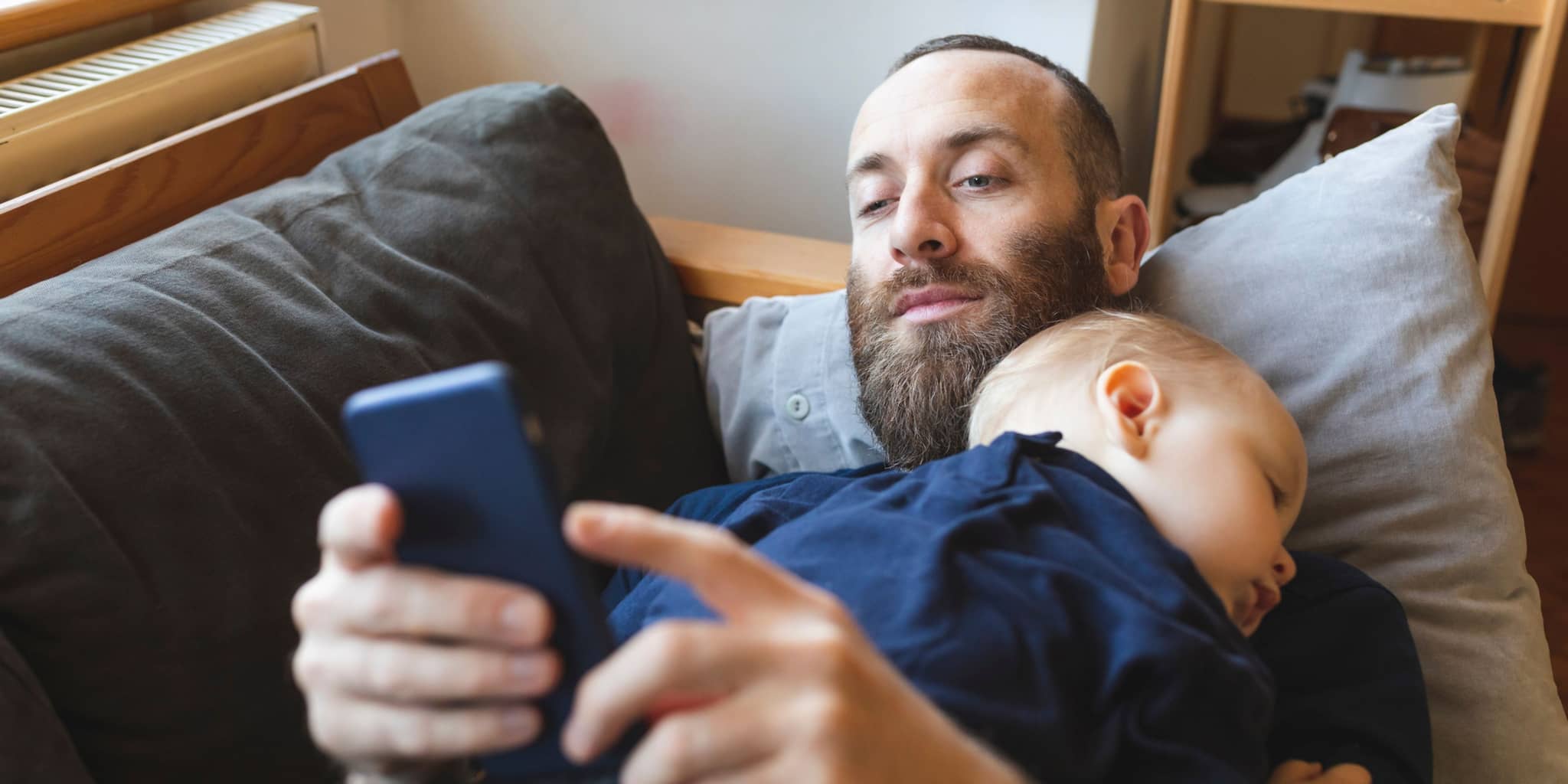 Ein Vater liegt mit seinem schlafenden Kind auf dem Sofa und schaut auf sein Handy.
