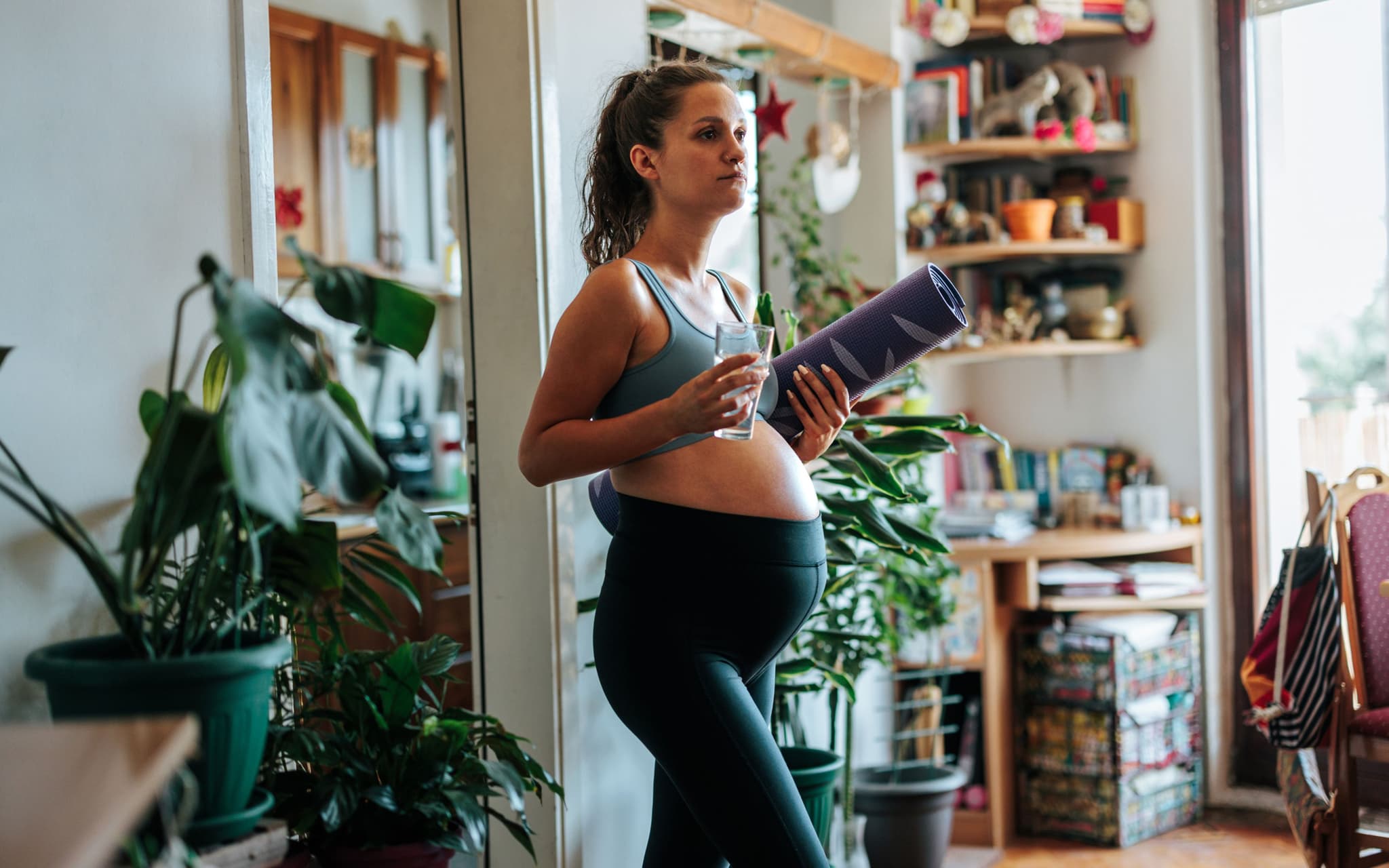 Schwangerschaftsmythen: Eine Schwangere Frau in Sportkleidung trägt eine Gymnastikmatte und trinkt ein Glas Wasser