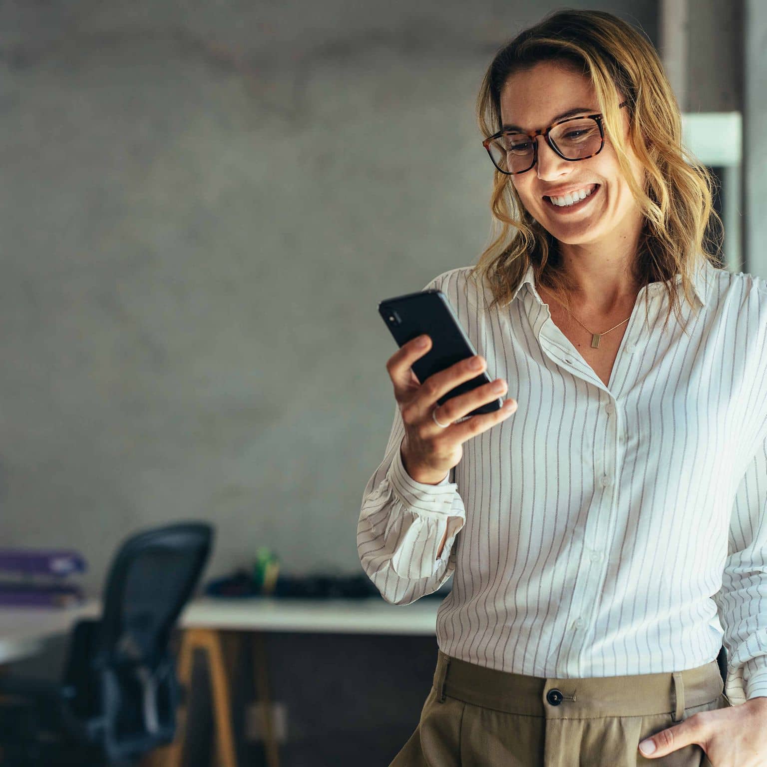 Image: Businesswoman standing in the office at ease, looking something up on her mobile phone.
