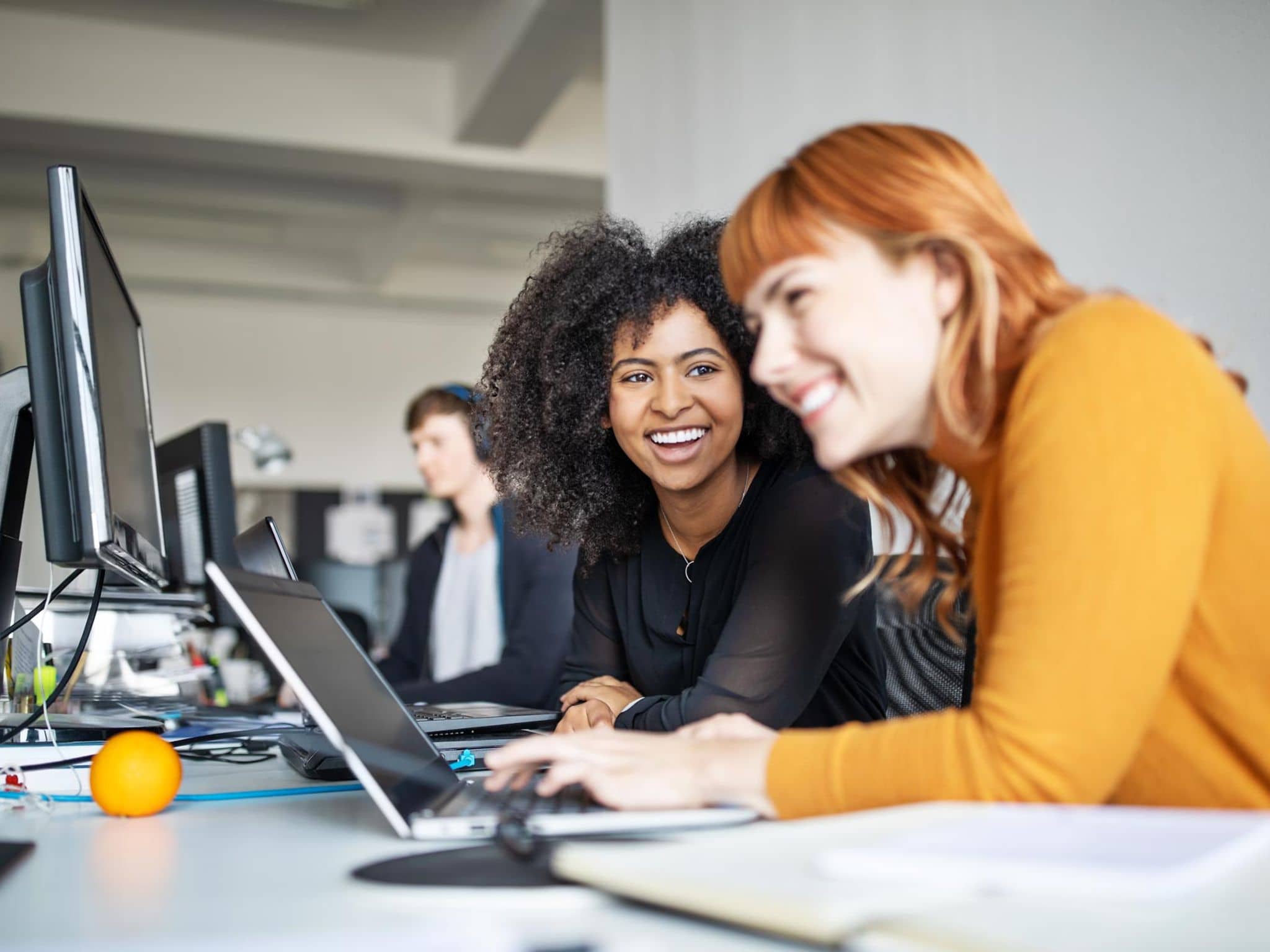 DAK Cashback 60: Zwei junge Frauen arbeiten in einem Großraumbüro zusammen an einem Computer.