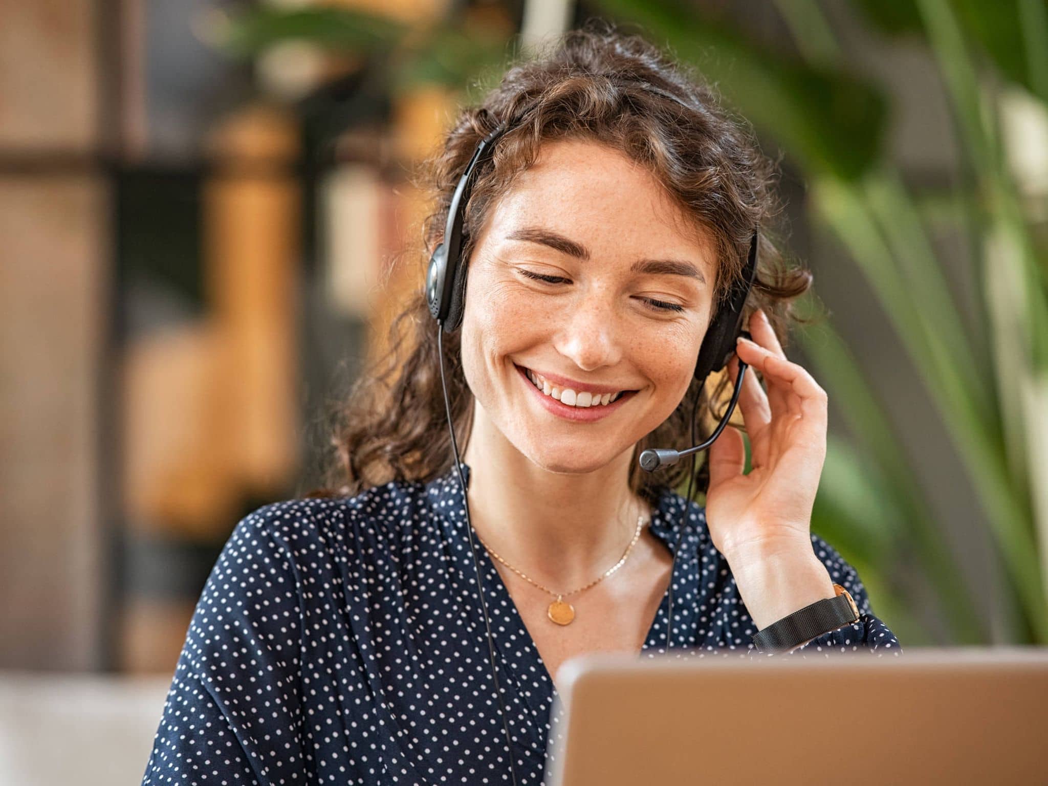 Lächelnde Frau mit Headset sitzt am Tisch und führt ein Gespräch, vermutlich im Kundenservice oder Callcenter.