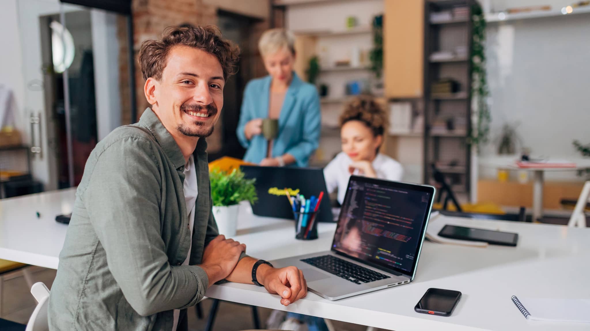 Nasmejani muškarac sedi sa kolegama za konferencijskim stolom ispred laptopa i gleda u kameru.